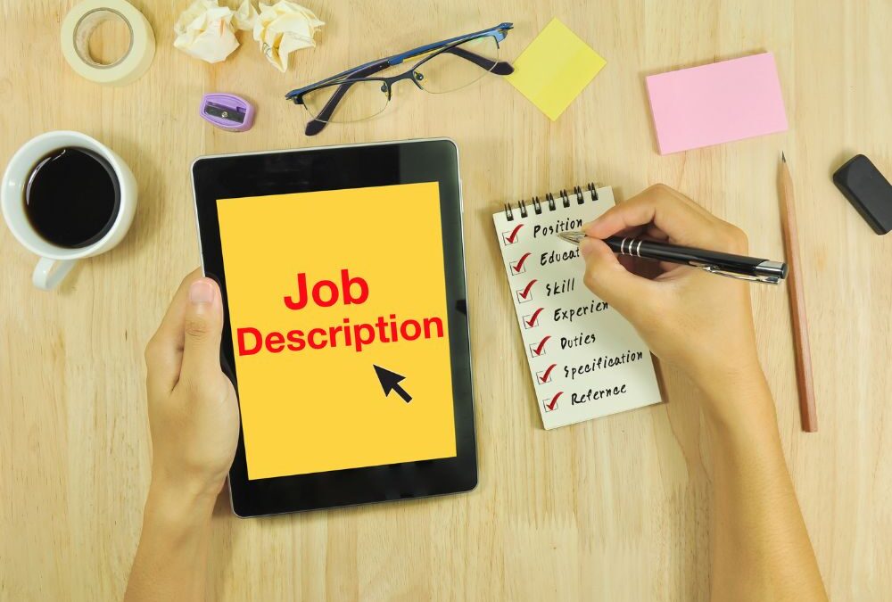 Hand holding a tablet with the words JobDescription displayed in red letters on a yellow background. The right hand holds a pen over a notepad and checks a list with the following items: position, education, skill, experience, duties, specification, reference.