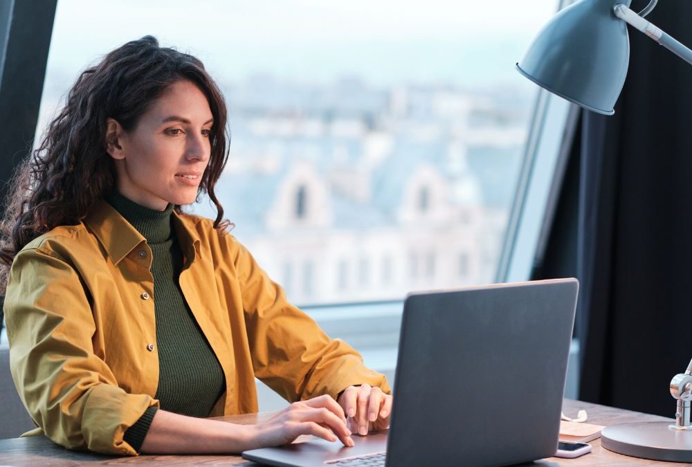 Young white woman in yellow shirt typing on a laptop in an office. There is a glass window on the background and you can see a blurried view of houses